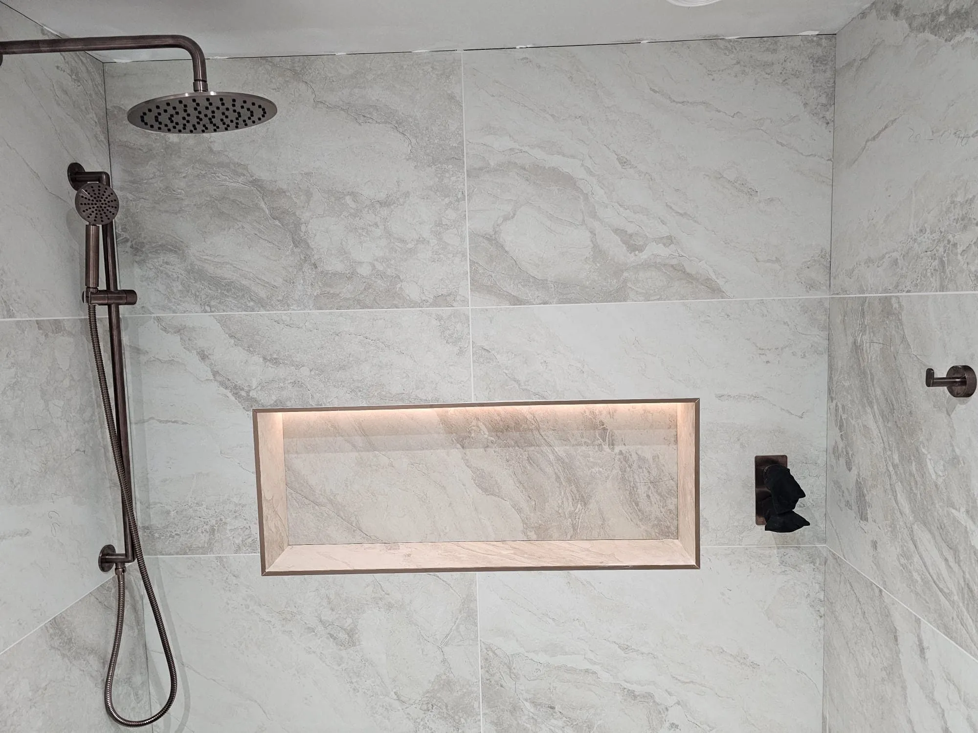 Walk-in shower with brushed bronze rainfall head, riser rail and lit storage niche on marble-look tiles