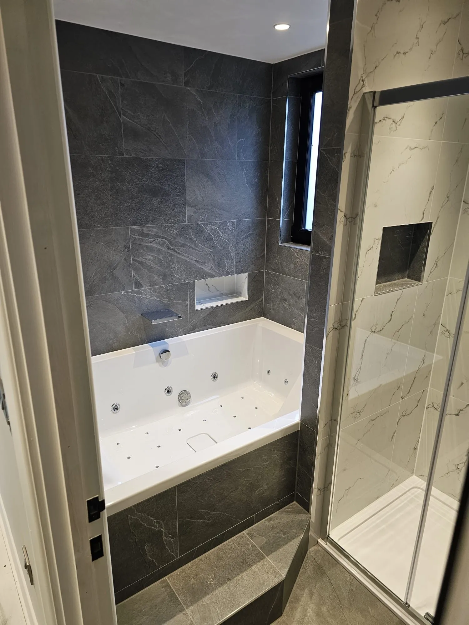 Fitted bath with shower screen beneath a skylight in a grey-tiled bathroom