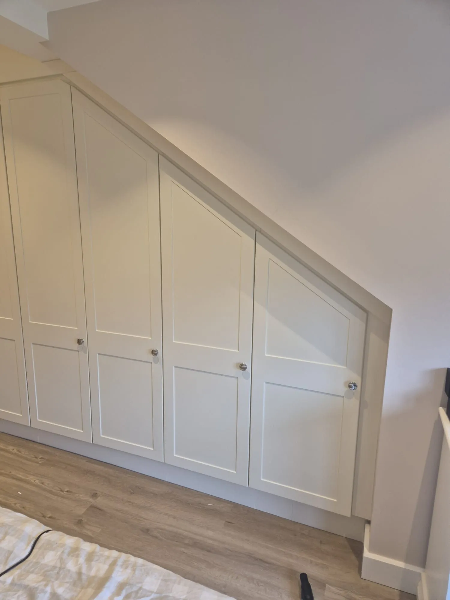 Fitted shaker wardrobes scribed to a sloping ceiling with chrome knobs and oak-effect floor