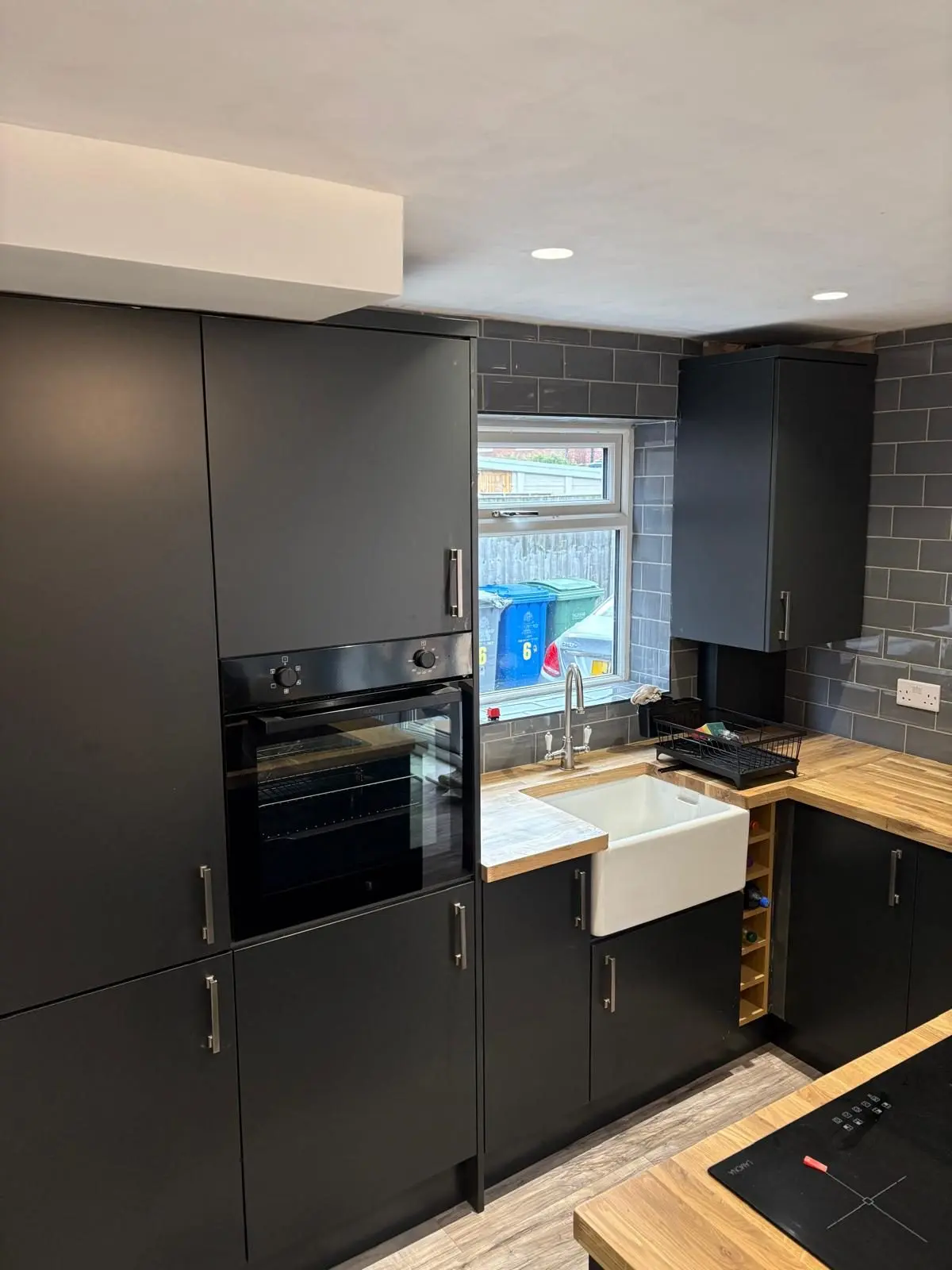 Tall black housing with integrated oven beside a Belfast sink and grey metro splashback